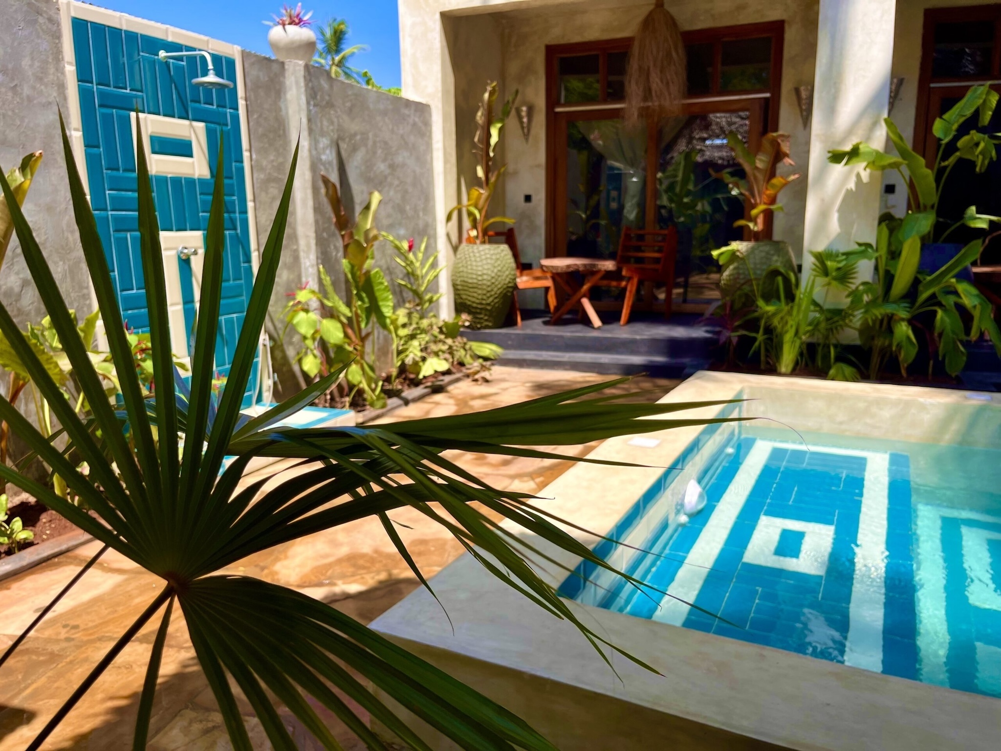 Sunny view of a private pool with a turquoise outdoor shower and tropical plants in a Zanzibar bungalow