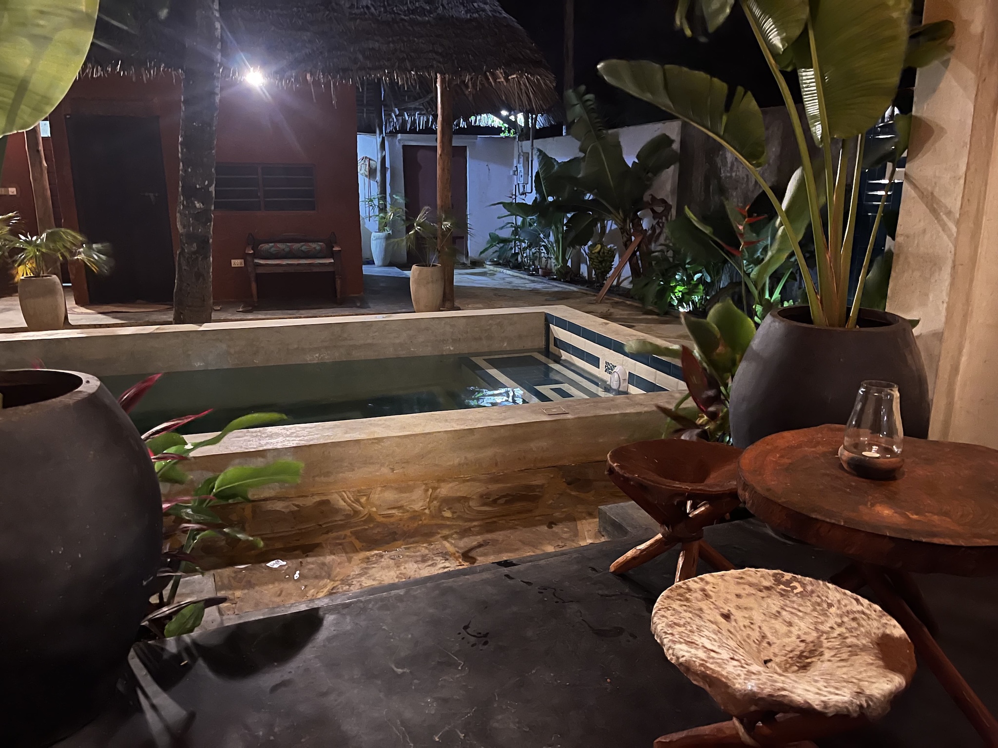 Private swimming pool at night in a Zanzibar bungalow courtyard with tropical plants and wooden furniture.